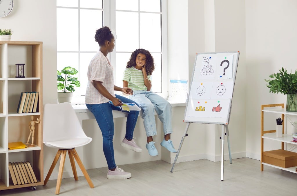 A child and therapist engaging in play therapy, drawing on a whiteboard together, smiling, bright and clinical but welcoming environment