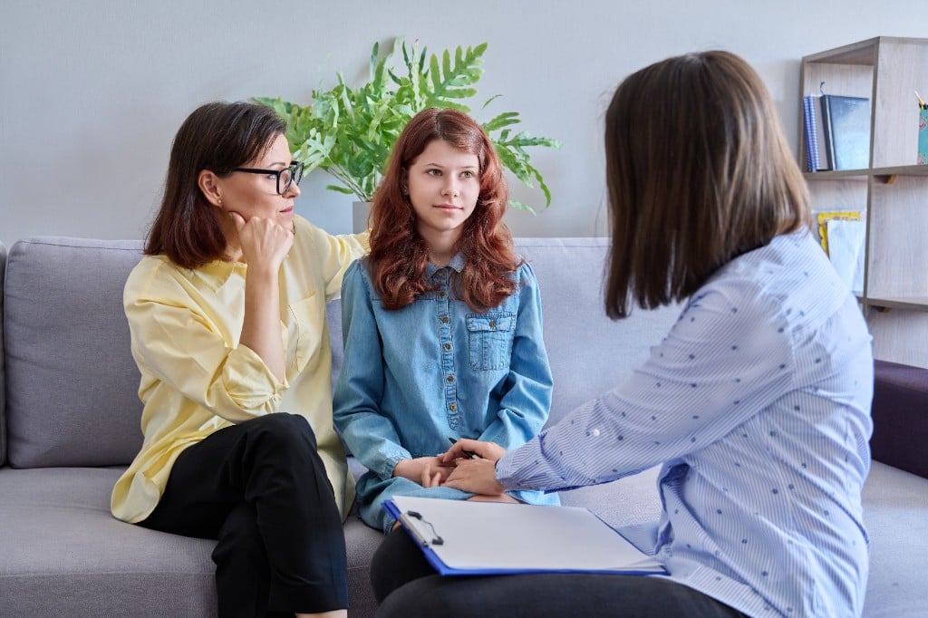 Two parents sitting together on a therapy couch, looking attentive and hopeful as they consult with a professional, warm and supportive atmosphere