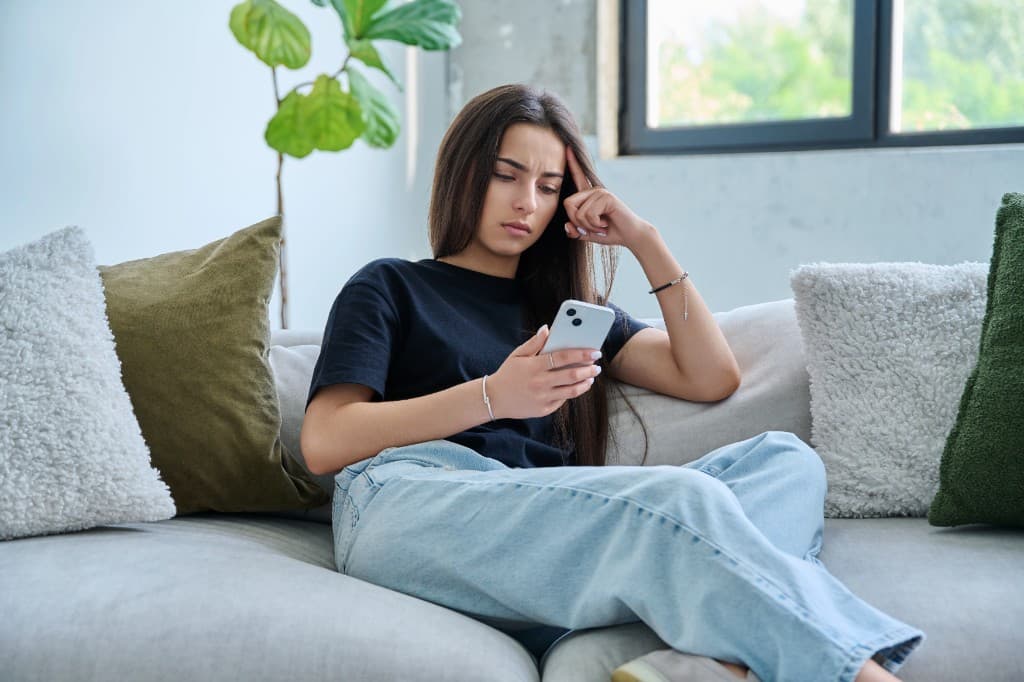 Teen sitting on a couch looking at their phone, worried and deep in thought.