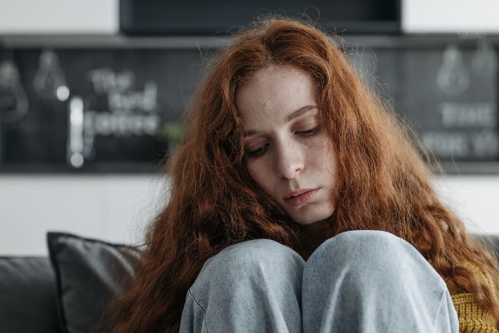 Withdrawn teenage girl with long red hair sitting on a couch hugging her knees and looking down, as a parent wonders whether it is teen depression or normal moodiness