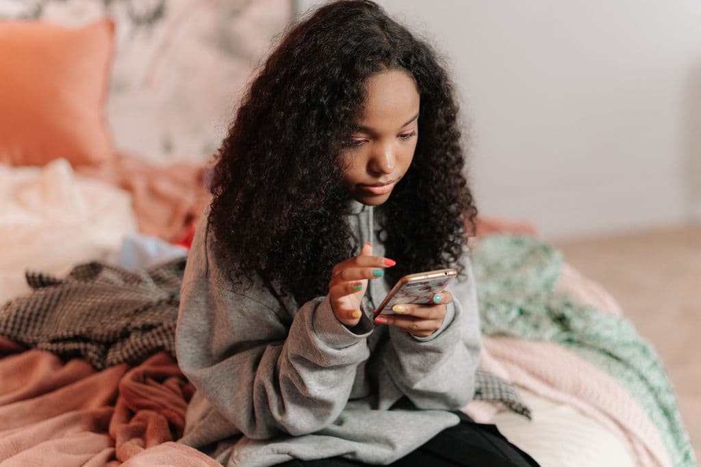 Teen girl sitting on her bed absorbed in her phone, illustrating everyday social media use and its effect on teen mental health