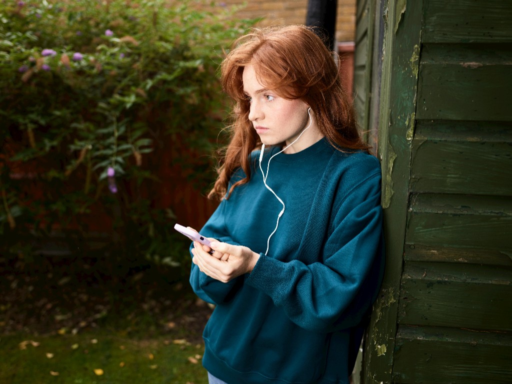 Teen girl in a teal sweatshirt with wired earbuds, looking pensive at her phone while leaning against a green wooden wall — illustrating quiet adolescent anxiety and emotional shutdown.
