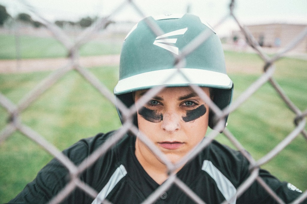 Teen baseball player in a helmet with eye black staring through a chain-link backstop before a game, illustrating signs of sports anxiety and pre-game pressure in young athletes