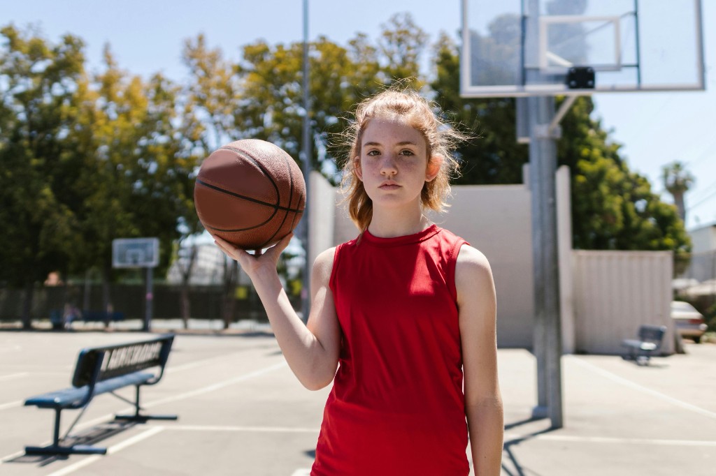 Teen girl in a red basketball jersey holding a ball on an outdoor court with a serious expression, illustrating youth sports pressure and the weight of athlete identity in adolescence