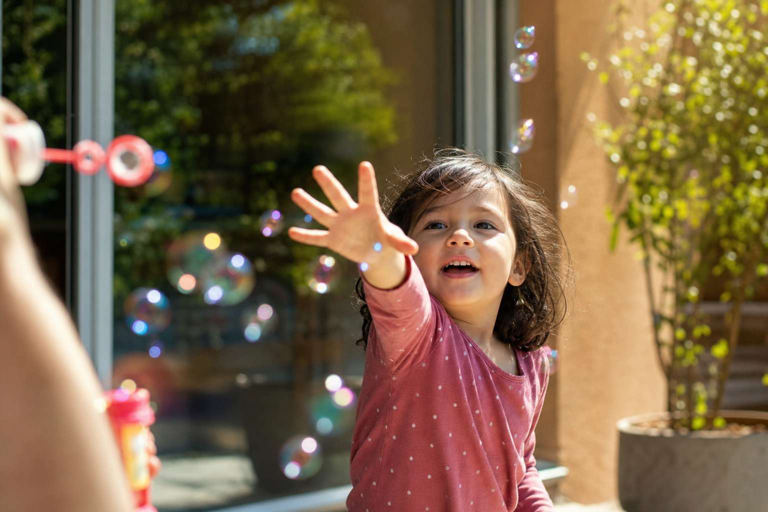 Young child reaching for floating soap bubbles outdoors while an adult holds a bubble wand, a real-life moment to practise a target word like more bubbles