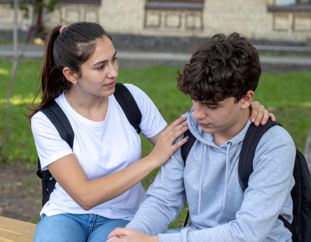 Teenage girl gently resting a hand on a withdrawn teen boy's shoulder while sitting together on an outdoor bench, illustrating a calm, low-pressure conversation with a teen who may be depressed