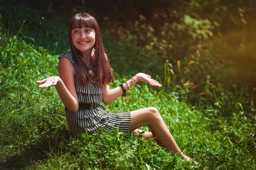 Relaxed, smiling teenage girl sitting outdoors on the grass and shrugging playfully, illustrating normal teenage ups and downs rather than depression