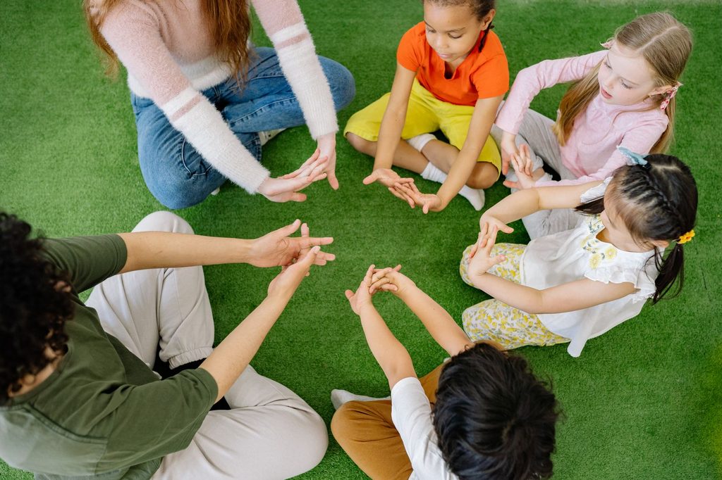 Children and instructors sitting in a circle on a green floor mat stretching their hands together, a fine motor warm-up used in pediatric occupational therapy