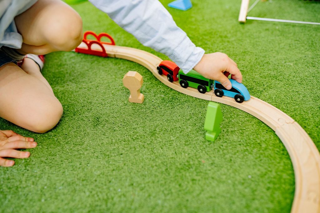 Young child guiding a wooden toy train along a curved track on a green play mat, building fine motor control that supports handwriting
