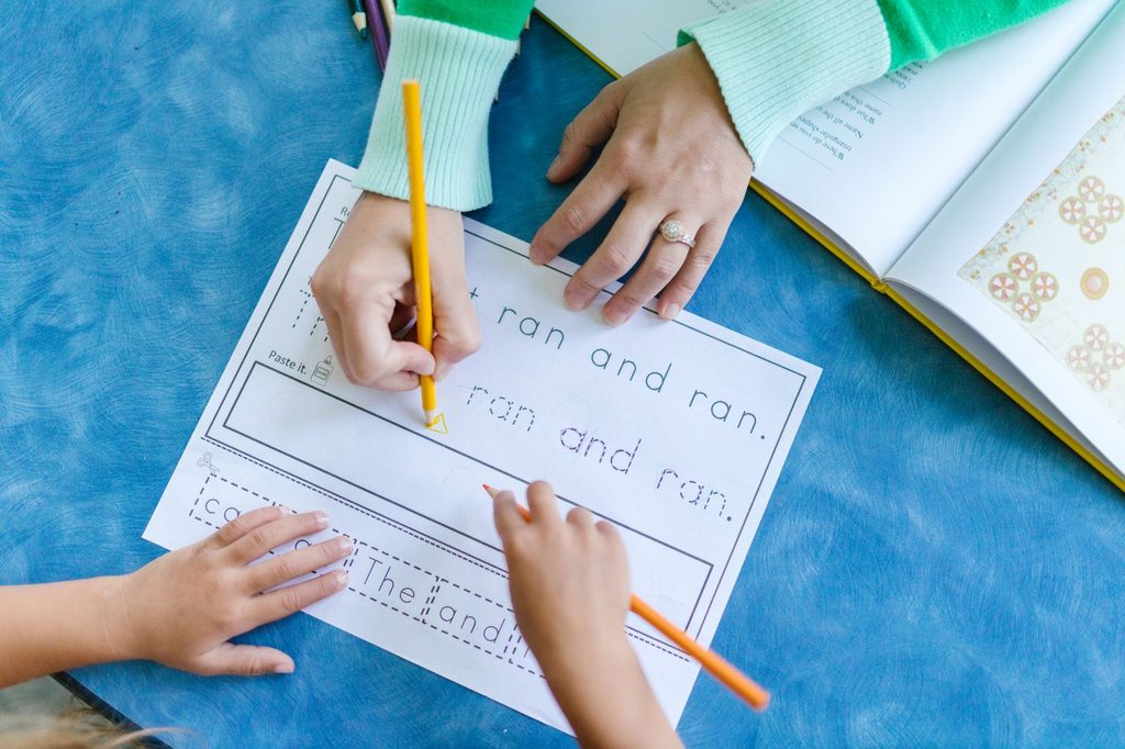 Child tracing and writing sentences with a yellow pencil while an adult guides a handwriting practice worksheet at a blue table